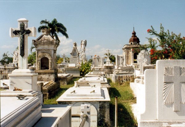 "Nachdem er seinen Touristendollar entrichtet, setzte Broschkus den Schritt in die Totenstadt, zögernd: linksrechts gewaltig umgitterte Schmucksarkophage, villenartig emporsäulende Gruften mit Steinkreuzen, die mitunter bis in die Maserung der Rinde und mit fein ausgemeißelten Jahresringen Holzkreuze imitierten ...": Santa Ifigenia